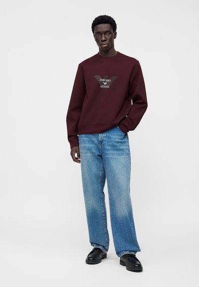 Άνδρας που φοράει μπλουζάκι Emporio Armani σε μαρονοκόκκινο χρώμα, ανοιχτόχρωμα μπλε τζιν και μαύρα παπούτσια, στέκεται με το ένα χέρι στην τσέπη σε λευκό φόντο.
