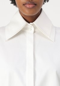 White shirt with a large, structured collar and two buttons at the neck, featuring a smooth, crisp fabric and subtle texture.