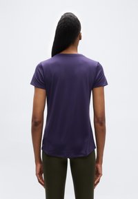 Γυναίκα με μακριά μαλλιά στέκεται με την πλάτη γυρισμένη, φορώντας ένα απλό μωβ κοντομάνικο μπλουζάκι και ελιά πράσινο παντελόνι σε λευκό φόντο.