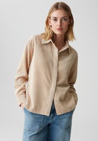 Camicia di velluto a coste beige con texture a righe verticali, chiusura frontale con bottoni e colletto a punta. Abbinata a pantaloni in denim blu larghi.