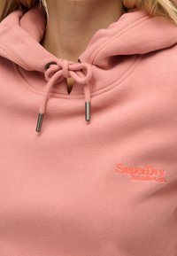 Sudadera rosa hecha de tela suave con capucha ajustable, que presenta un logo bordado de "SuperDry" en un color a juego en el pecho.