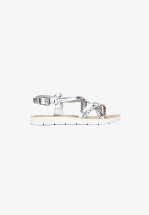 Sandalias planas metálicas plateadas con correas trenzadas, una hebilla ajustable en el tobillo y una suela de goma blanca. Textura suave y diseño minimalista.