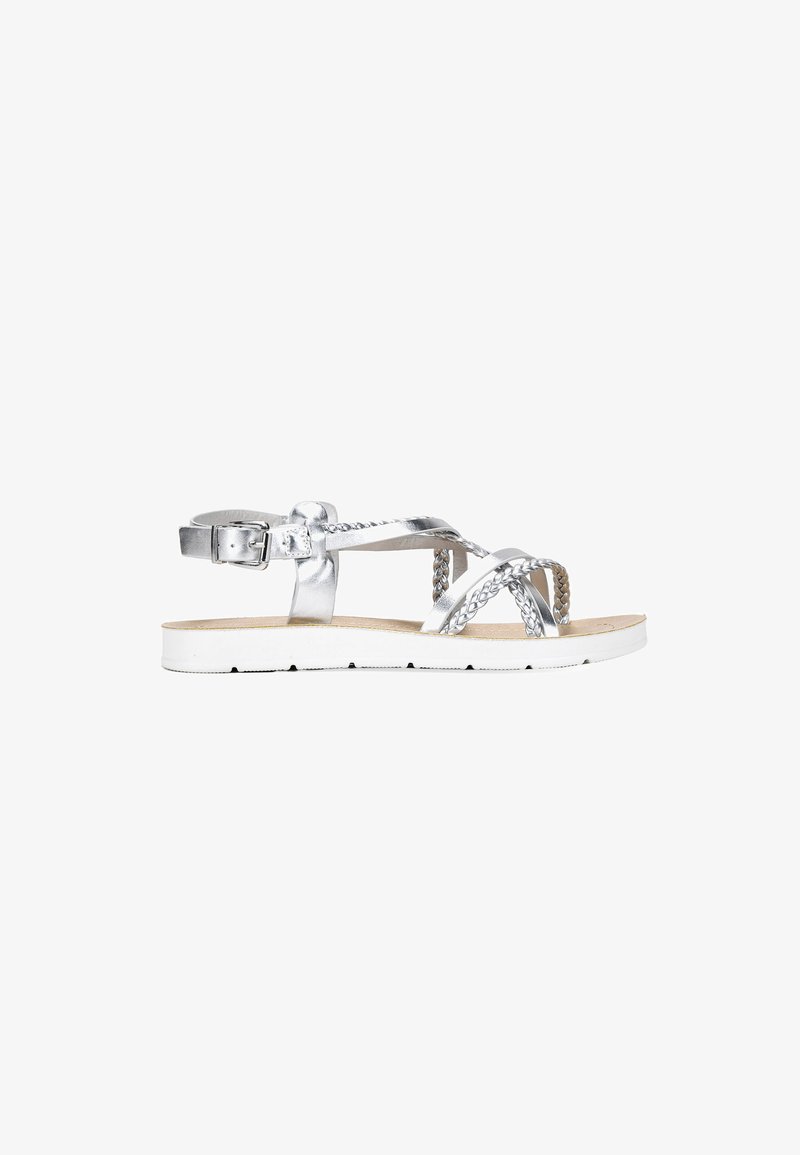 Sandalias planas metálicas plateadas con correas trenzadas, una hebilla ajustable en el tobillo y una suela de goma blanca. Textura suave y diseño minimalista.