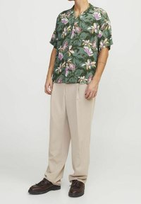 Kortärmad grön skjorta med rosa och vita blommönster, tillsammans med ljusbeiga byxor och mörka skor, stående på en neutral bakgrund.