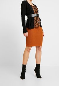 Cárdigan negro de canalé superpuesto sobre una blusa a rayas naranja y una falda naranja ajustada con un acabado texturizado; combinado con botas de tacón negras hasta el tobillo.