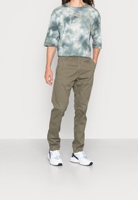 Pánské tričko s krátkým rukávem v modrých a zelených odstínech s vazbou barvy, doplněné olivově zelenými zúženými kalhotami a bílými teniskami s námořnickými akcenty.