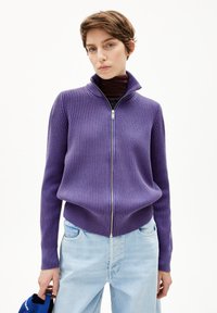 Lila gerippter Pullover mit hohem Kragen und Reißverschluss, kombiniert mit hellblauen Jeans. Der Stoff wirkt weich und strukturiert.