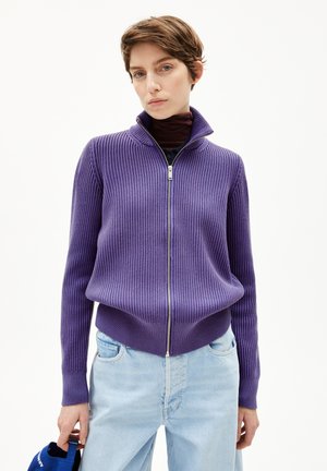 Maglione viola a coste con colletto alto e chiusura con zip, abbinato a jeans azzurri chiari. Il tessuto appare morbido e testurizzato.