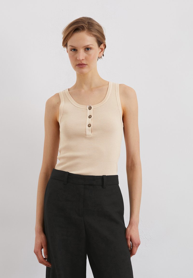 Débardeur beige à côtes avec un col en rond et trois boutons marron sur le devant, associé à un pantalon noir taille haute et ajusté.