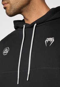 Camisola preta com capuz e cordões cinza claro, com um logótipo gráfico prateado e um desenho de cobra na frente, feita de um tecido macio.