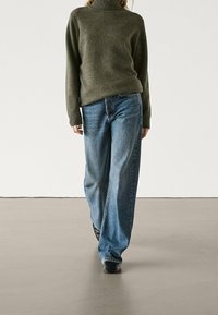 Person wearing olive green turtleneck sweater, loose blue jeans, and black shoes standing on a smooth floor against plain white wall.