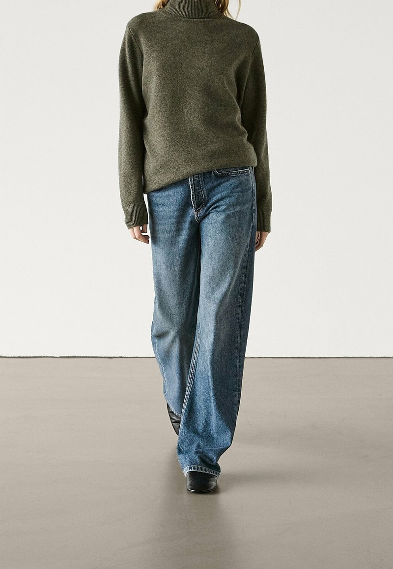 Person wearing olive green turtleneck sweater, loose blue jeans, and black shoes standing on a smooth floor against plain white wall.