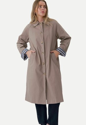 Femme portant un manteau beige boutonné avec des poignets à carreaux, mains dans les poches, regardant sur le côté, assorti à un pantalon foncé.