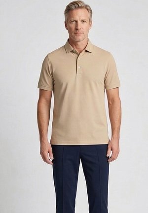 Blonde man met een beige poloshirt met korte mouwen en donkerblauwe broek, staand tegen een effen lichte achtergrond.
