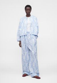 Mujer con chaqueta suelta de color azul claro con estampado floral y pantalones anchos a juego, con camiseta blanca y zapatos planos granate.