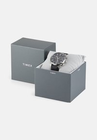 Montre Timex dans une boîte grise, avec un cadran noir, des accents argentés, des fonctions chronographe et un bracelet en cuir reposant sur un coussin gris.