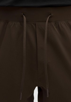 Elastický pas s dlouhými hnědými šňůrkami na tmavě hnědých měkkých kalhotách nebo kraťasech, zobrazující švy a nasbíraný materiál.