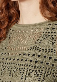 Olivgrüner Stricktop mit Lochmuster, das ein geometrisches Muster zeigt und eine schwarze Unterlage, die durch die Löcher sichtbar ist.