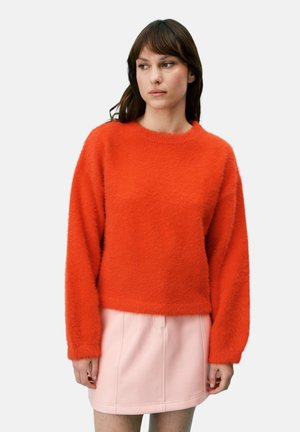 Femme aux cheveux bruns portant un pull pelucheux orange vif et une mini-jupe rose clair, regardant sur le côté contre un fond blanc.