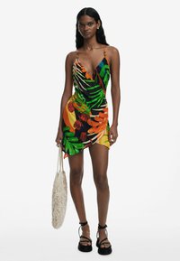 Vestido envolvente colorido con un patrón de hojas y flores tropicales, hecho de una tela ligera, con tirantes finos y un dobladillo diagonal.