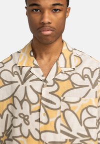 Joven con una camisa de botones de patrón floral en beis y amarillo, con collar y bolsillo en el pecho.