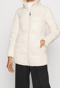 Persona che indossa un giubbotto imbottito beige chiaro con cappuccio, chiuso con la zip, con una mano in tasca, abbinato a pantaloni neri larghi.