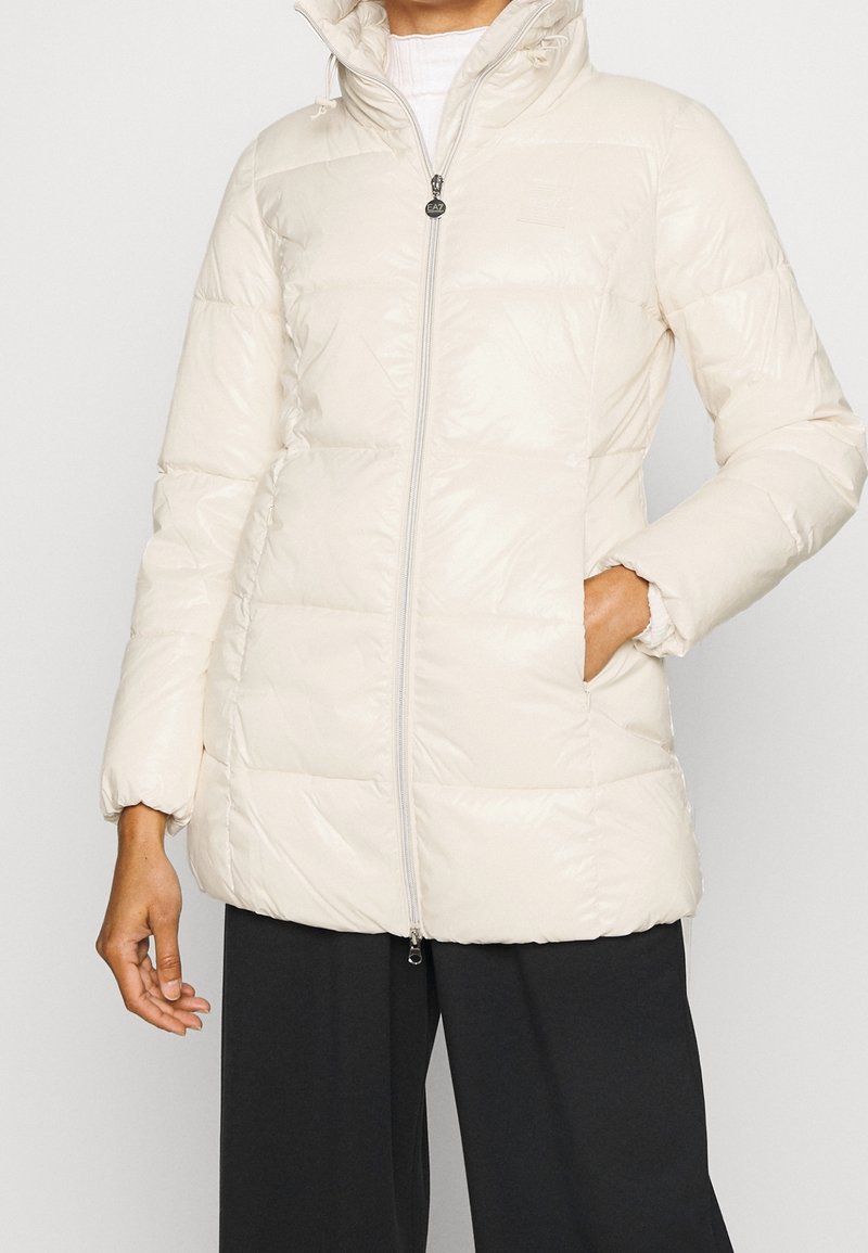 Persona che indossa un giubbotto imbottito beige chiaro con cappuccio, chiuso con la zip, con una mano in tasca, abbinato a pantaloni neri larghi.