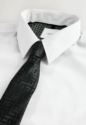 Valkoinen paitapusero, jossa on jämäkkä kaulus, yhdistettynä mustaan solmuun, jossa on geometrinen kuvio. Kauluksessa on kuusikulmainen kuvio.