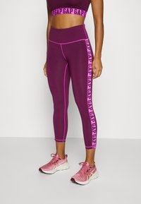 Lila träningsset bestående av en kort topp och leggings med rosa detaljer och logotyp längs sidan, tillverkat av elastiskt material.