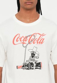 Valge puuvillane t-särk, millel on graafiline disain kauboiharul, punane Coca-Cola logo ja joonistuslikud mustad ja valged aktsendid.