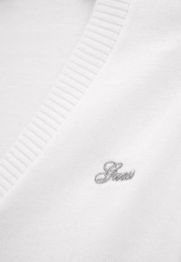 Pull en tricot blanc avec un col en V, présentant un côtelé texturé au niveau du col. Petit logo brodé gris en bas à gauche.