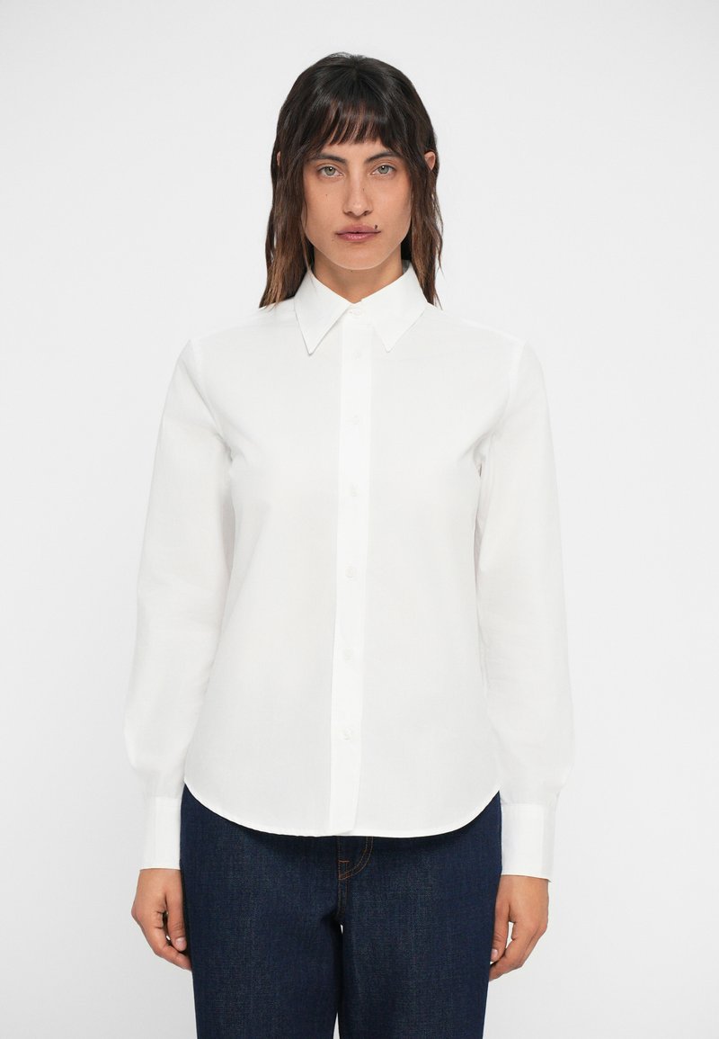 Camicia bianca a maniche lunghe con colletto, chiusura frontale a bottoni e orlo arrotondato. Tessuto liscio e vestibilità su misura con maniche con polsini.
