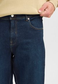 Jeans de mezclilla azul oscuro con un diseño clásico de cinco bolsillos, cierre de botón y costuras naranjas en contraste. Presentan una textura suave.