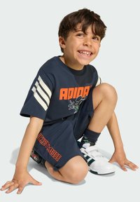 Maglietta Adidas blu navy con logo e grafica arancione, abbinata a pantaloni corti coordinati, sneaker bianche e verdi e calze scure.