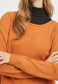 Oranger Wollpullover mit Rundhalsausschnitt, gerippten Bündchen und weicher Textur, über einem schwarzen Rollkragenpullover getragen. Sichtbare Nahtdetails.