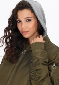 Giacca bomber verde oliva con cappuccio grigio, chiusura con zip, polsi a costine e tasche laterali. Tessuto texturizzato con maniche arricciate.