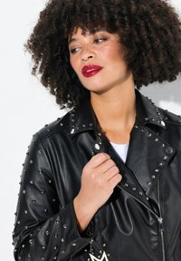 Black leather jacket with silver studs, featuring a classic collar and zipper closure. Model holds the jacket slightly closed, revealing a white top.