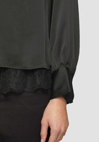 Zwarte blouse met geplooide stof, kantafwerking aan de zoom en satijnachtige mouwen. Close-up van de arm en stofdetails.