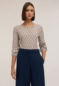 Top beige con motivo geometrico abbinato a pantaloni a gamba larga navy. Il top presenta una stampa geometrica e un colletto con gioielli. Maniche lunghe arrotolate fino a metà avambraccio.