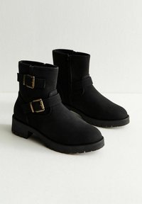 Para czarnych botków za kostkę z bocznymi zamkami, dekoracyjnymi podwójnymi paskami i metalowymi klamrami na jasnym tle.