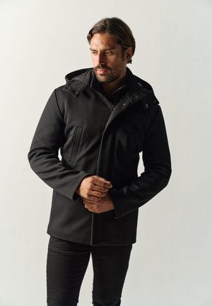 Mann trägt eine schwarze Kapuzenjacke und schwarze Hose, steht mit gefalteten Händen und schaut leicht zur Seite vor einem schlichten hellen Hintergrund.