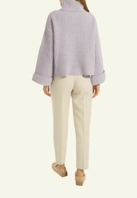 Helllila, übergroßer Strickpullover mit hohem Kragen, geripptem Muster und weiten Ärmeln, kombiniert mit cremefarbenen schmal zulaufenden Hosen und beigen Clogs.