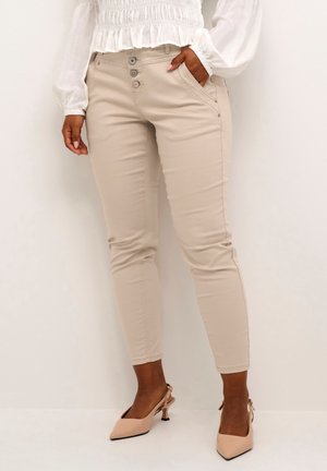 Kobieta ubrana w beżowe, dopasowane spodnie z trzema guzikami oraz białą, długorękawową bluzkę, stojąca na jednolitym jasnym tle.