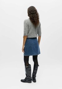 Femme aux cheveux bouclés portant un pull gris, une jupe en jean bleue, des collants noirs et des bottes noires montantes, debout dos à la caméra.
