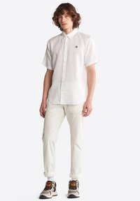 Camisa blanca de manga corta con botones, confeccionada en una tela ligera, presenta un pequeño logo bordado en el pecho, combinada con pantalones color crema y zapatillas.