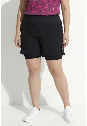 Persona con pantalones cortos negros en capas, una camiseta rosa con estampado y zapatillas blancas, de pie contra un fondo claro y liso.