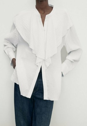 Personne portant un blouse blanche texturée avec un grand col superposé et un jean bleu foncé, se tenant les mains dans les poches contre un fond blanc.