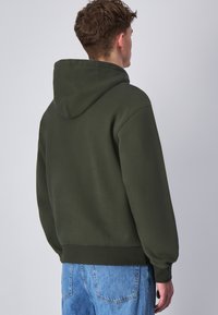 Felpa verde scuro con cappuccio arrotondato, polsini e orlo a coste, realizzata in materiale morbido. Indossata con jeans in denim azzurri.