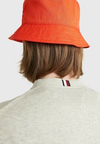 Gorra de cubo naranja utilizada sobre un sudadera gris claro, con un cuello acanalado y un pequeño detalle de franjas tricolores en la parte de atrás.