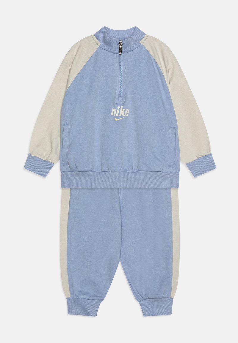 Lichtblauwe Nike trainingspak met een col met halve rits, crèmekleurige accentmouwen, elastische manchetten en elastische tailleband. Bevat logo op de borst.